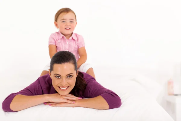 Lycklig liten flicka sitter på mammas rygg på sängen — Stockfoto
