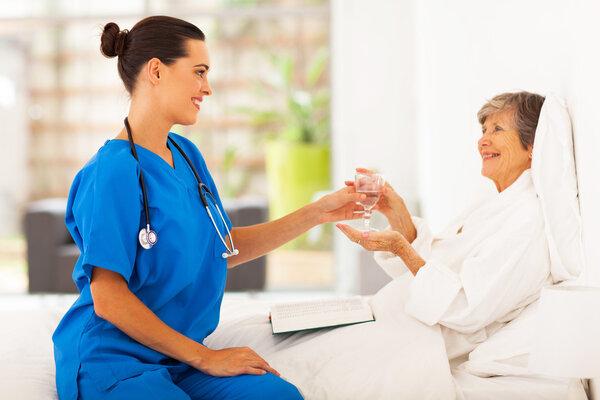 Caring young caregiver bring a glass of water to senior woman on bed