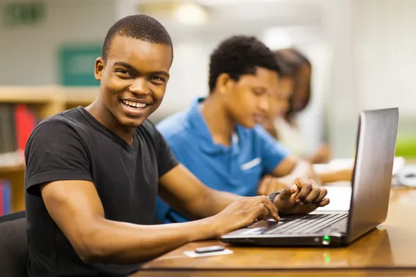 Feliz estudante universitário africano masculino usando laptop na biblioteca — Fotografia de Stock
