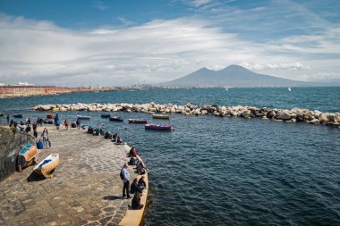 Sakin mavi Tyrhenian Denizi. Napoli 'nin setinden Vezüv Dağı yanardağına bakın. Taştan dalgakıranların yakınına demirlemiş eğlence tekneleri. İnsanlar izin günlerinde rahatlar ve sahil boyunca yürürler..
