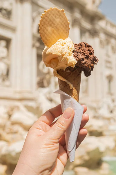 Gelato is Italian ice cream. Ice cream cone in a woman's hand against the backdrop of the Trevi Fountain. Chocolate vanilla ice cream with a waffle circle.