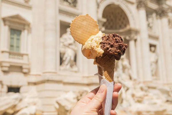 Gelato is Italian ice cream. Ice cream cone in a woman\'s hand against the backdrop of the Trevi Fountain. Chocolate vanilla ice cream with a waffle circle.