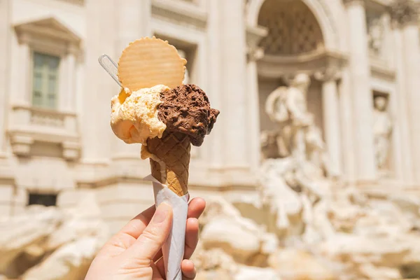 Gelato is Italian ice cream. Ice cream cone in a woman\'s hand against the backdrop of the Trevi Fountain. Chocolate vanilla ice cream with a waffle circle.
