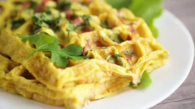 Yeşillikle doldurulmuş yumurta omleti ve waffle şeklinde kızarmış sosis, ahşap bir masada.