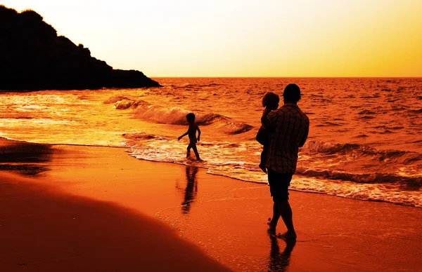 Pai e duas crianças silhuetas na praia ao pôr do sol — Fotografia de Stock