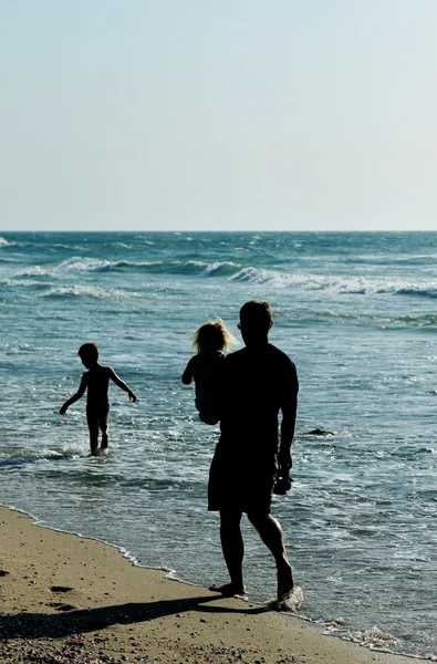 Pai e duas crianças silhuetas na praia ao pôr do sol — Fotografia de Stock