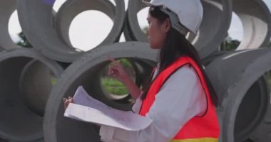 Beyaz bir miğferdeki kadın mühendis bir plandaki verileri okuyor ve inşaat alanındaki su sistemini kontrol ediyor.