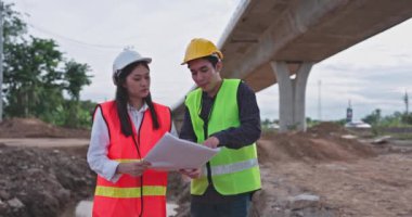 Güvenlik önlüğü ve kask takan Asyalı genç inşaat mühendisleri, yapının bir planını ellerinde tutarak inşaat altındaki bir otoyolda duruyorlar ve.