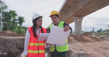 Güvenlik önlüğü ve kask takan Asyalı genç inşaat mühendisleri, yapının bir planını ellerinde tutarak inşaat altındaki bir otoyolda duruyorlar ve.