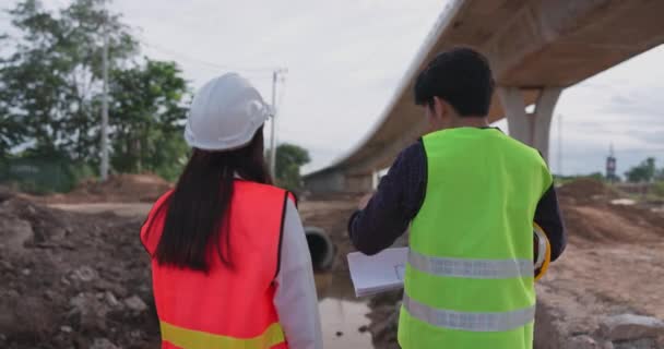 安全ジャケットとヘルメットを身に着けている若いアジアの土木技術者は 建設中の高速道路に立ち 構造の青写真を保持し 議論する — ストック動画