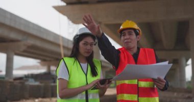 Güvenlik yeleği ve kask giyen iki Asyalı mühendis inşaat sahasındaki kablosuz tablet bilgisayardaki bir inşaat planlayıcısına bakarken bir projeyi tartışıyorlar..