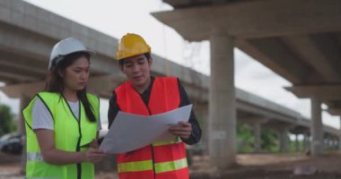 Güvenlik yeleği ve kask giyen iki Asyalı mühendis inşaat sahasındaki kablosuz tablet bilgisayardaki bir inşaat planlayıcısına bakarken bir projeyi tartışıyorlar..