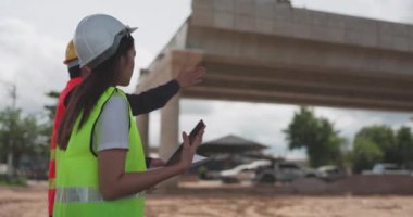Güvenlik yeleği ve kask giyen iki Asyalı mühendis inşaat sahasındaki kablosuz tablet bilgisayardaki bir inşaat planlayıcısına bakarken bir projeyi tartışıyorlar..