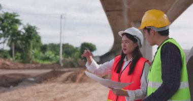 Güvenlik yeleği ve kask giyen iki Asyalı mühendis inşaat sahasındaki kablosuz tablet bilgisayardaki bir inşaat planlayıcısına bakarken bir projeyi tartışıyorlar..