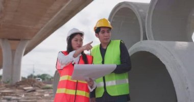 Güvenlik yeleği ve kask giyen iki Asyalı mühendis inşaat sahasındaki kablosuz tablet bilgisayardaki bir inşaat planlayıcısına bakarken bir projeyi tartışıyorlar..