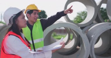 Güvenlik yeleği ve kask giyen iki Asyalı mühendis inşaat sahasındaki kablosuz tablet bilgisayardaki bir inşaat planlayıcısına bakarken bir projeyi tartışıyorlar..