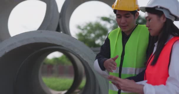 安全ベストとヘルメットを身に着けている2人のアジアのエンジニアの従業員は 建設現場のワイヤレスタブレットコンピュータ上の建設プランナーを見ている間 プロジェクトについて議論します — ストック動画