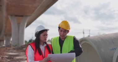Güvenlik yeleği ve kask giyen iki Asyalı mühendis inşaat sahasındaki kablosuz tablet bilgisayardaki bir inşaat planlayıcısına bakarken bir projeyi tartışıyorlar..