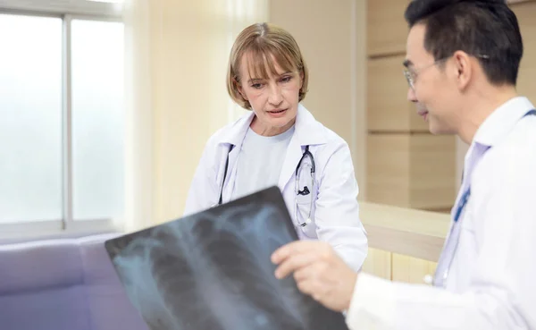 X-ray prints are examined by hospital staff. While looking at an x-ray image, two male medics confer. Two Caucasian doctors examine an MRI image and discuss it.