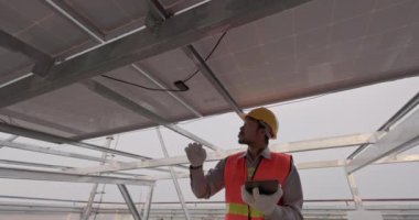 Adam tablet ekranı ve güneş panelini yakından kontrol ediyor. Sert bir kask takan ve üniforma giyen bir adam yenilenebilir enerji çiftliğinde durur ve bir cihaz çalıştırır..
