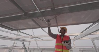 Mühendislerden biri tablet ekranı ve güneş panelini yakından kontrol ediyor. Sert bir kask takan ve üniforma giyen bir adam yenilenebilir enerji çiftliğinde durur ve bir cihaz çalıştırır..