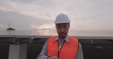 Mühendislerden biri tablet ekranı ve güneş panelini yakından kontrol ediyor. Sert bir kask takan ve üniforma giyen bir adam yenilenebilir enerji çiftliğinde durur ve bir cihaz çalıştırır..