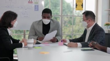 Bir konferans masasında, yüz maskesi takan çok ırklı erkek ve kadın yatırımcılardan oluşan bir grup iş belgelerini tartışıyor..