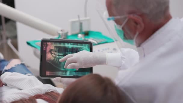 在当代的一家牙科诊所 一位戴着面具的白人男牙医用平板电脑给一位快乐的女病人提供了如何恢复牙齿的建议 — 图库视频影像