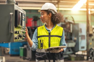 Kontrol panelinin önünde duran kadın bir endüstriyel elektrik mühendisi kafasında güvenlik başlığı ve elinde tabletle bir fabrikada CNC makinelerini kontrol ediyor ve koruyor..