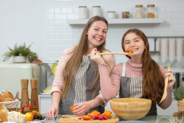 Beyaz, lekesiz, modern bir mutfakta, gülümseyen bir anne ve kız akşam yemeği için sebzeleri nasıl hazırlayacaklarını gösteriyorlar..