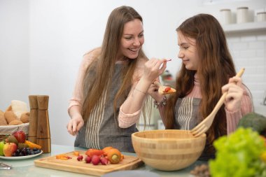 Beyaz, lekesiz, modern bir mutfakta, gülümseyen bir anne ve kız akşam yemeği için sebzeleri nasıl hazırlayacaklarını gösteriyorlar..