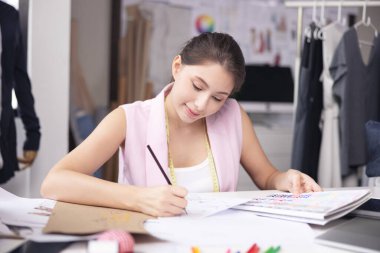 Bir atölyede, gülümseyen Asyalı moda tasarımcısı bir dizüstü bilgisayarda çalışıyor..