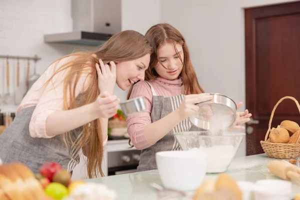 Mom and her younger kids daughter in a modern kitchen area celebrating joyous learning, baking, cake decoration, happy small daughter make prepare sweet pastries with caring mom on weekend at home