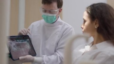 Genç bir dişçi, dişçi koltuğundaki kadın hastaya dijital tablette röntgen gösteriyor..