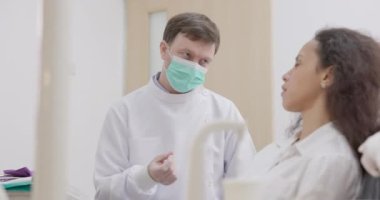 Koltuktaki dişçi kadın erkek dişçiyle dişçi sandalyesindeki dişçi kadınla konuşuyor.