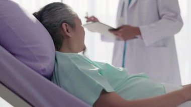 Asyalı bir erkek doktor semptomları inceler ve hastane yatağında yaşlı bir hastayla konuşur..