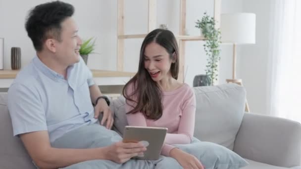 アジア系のカップル不幸な男はデジタルタブレットを保持しながら ソファの上にタブレットを使用して彼のパートナーを見ています 4K解像度 — ストック動画
