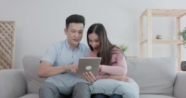 Birkaç Asya kökenli mutsuz adam, elinde dijital tabletle kanepede oturan ortağına bakıyor. 4K çözünürlüğü