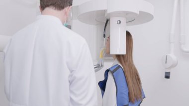 Panoramik röntgen makinesinde dişçi bir kadının ısırığını hizalar. Bir diş kliniğinde, kurşun önlük giymiş bir kadın kafasının etrafında dönen panoramik bir röntgen makinesi gibi hareketsiz durur.