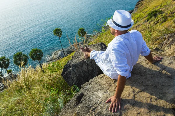 The man in a white suit and hat sitting on a rock on the sea ba — Stock Photo, Image