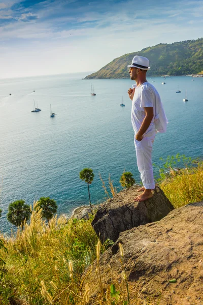 The man in a white suit and hat sitting on a rock on the sea ba — Stock Photo, Image