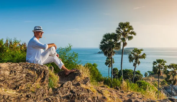 The man in a white suit and hat sitting on a rock on the sea ba — Stock Photo, Image