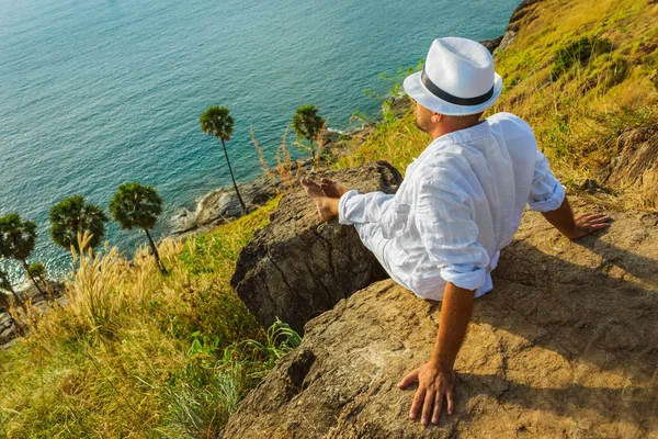 The man in a white suit and hat sitting on a rock on the sea ba — Stock Photo, Image