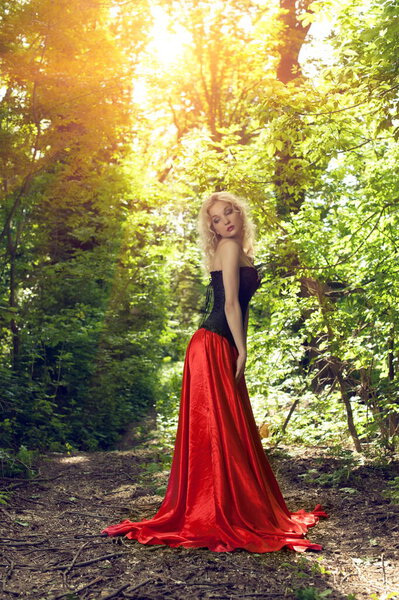 Beautiful sexy blonde in a black corset and red skirt with a long train posing in forest