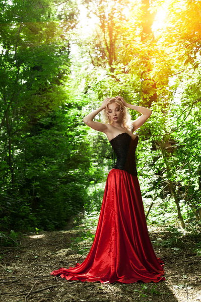 Beautiful sexy blonde in a black corset and red skirt with a long train posing in forest