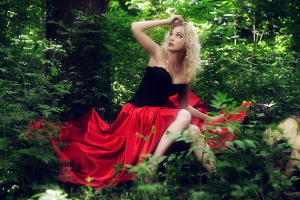 Beautiful sexy blonde in a black corset and red skirt with a long train posing sitting in green foliage