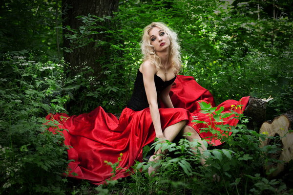 Beautiful sexy blonde in a black corset and red skirt with a train length posing in a forest in the green foliage