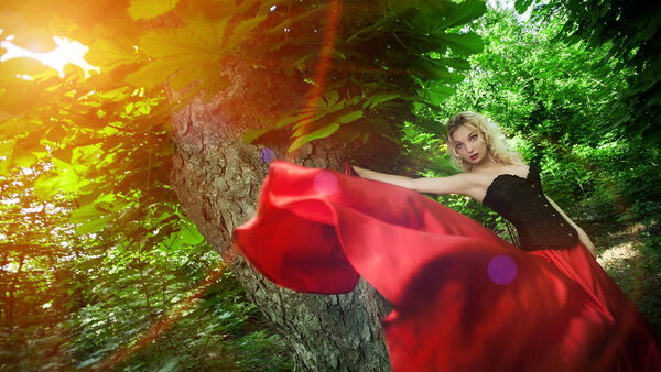 Beautiful sexy blonde girl in a black corset and red skirt with a long train posing in green leaves at the tree