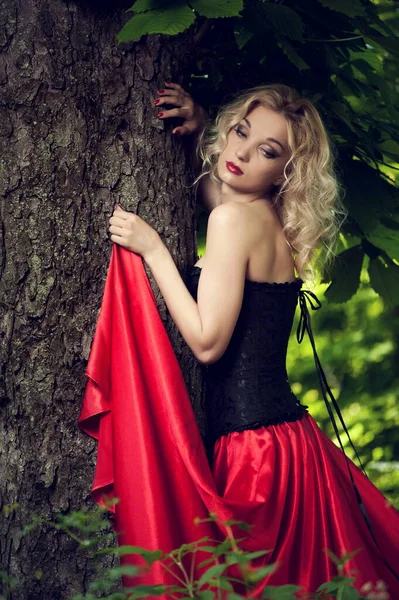 身穿黑色紧身胸衣和红色裙子 坐火车 身穿绿叶长裙的漂亮性感金发姑娘站在树旁 — 图库照片