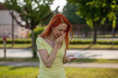 Alerjinin sert sadeleri. Genç kızıl saçlı kadın çiçek açan ağacın önünde hapşırıyor. Polen alerjisi belirtileri